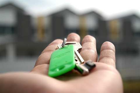 Outstrectched hand with a set of keys on a green keychain pointing towards house in the distance